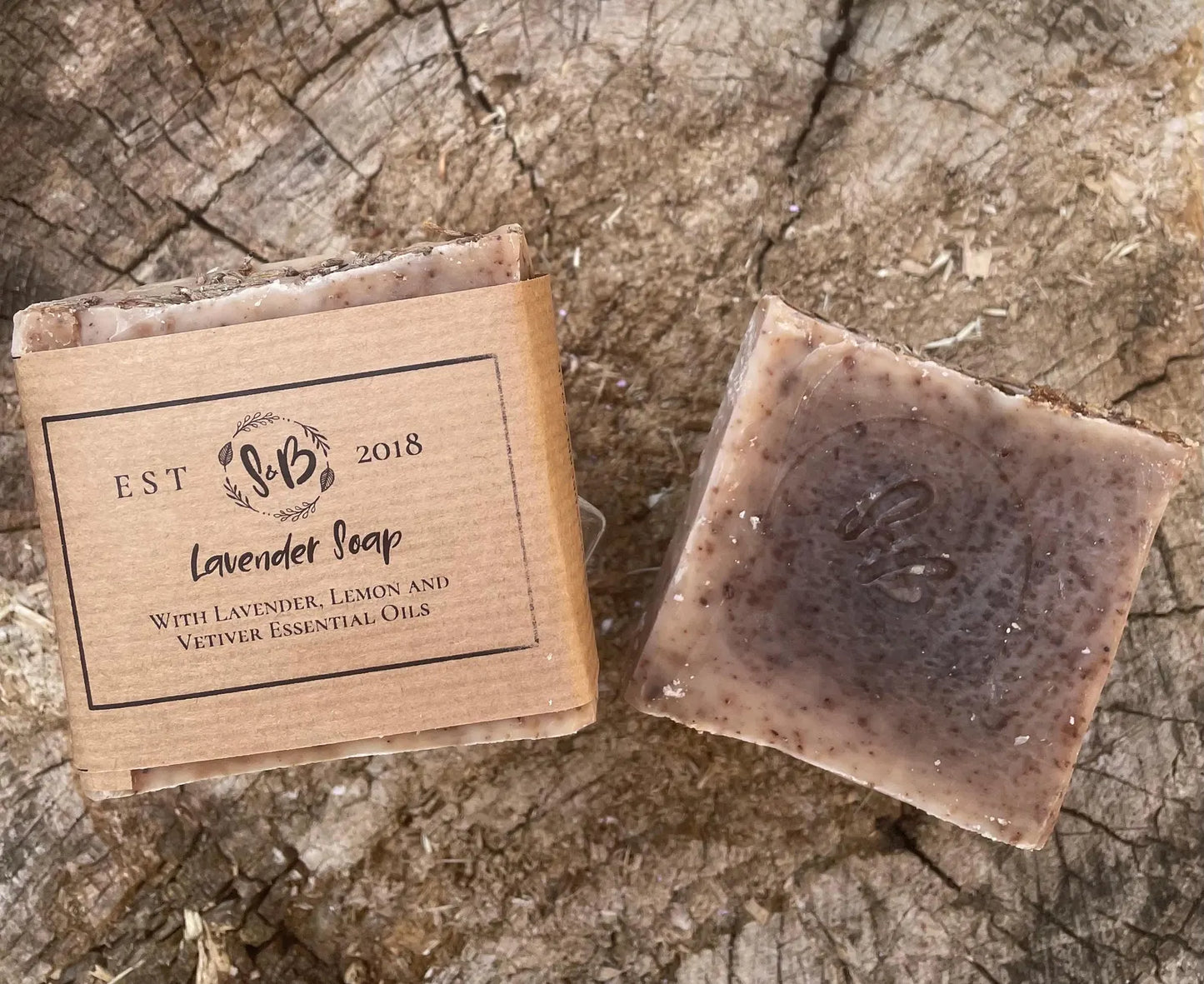 Handmade Lavender Soap bar by Soapy & Bubbly, unwrapped to show the rich texture and S&B stamp, next to its kraft paper packaging on a rustic wood background.
