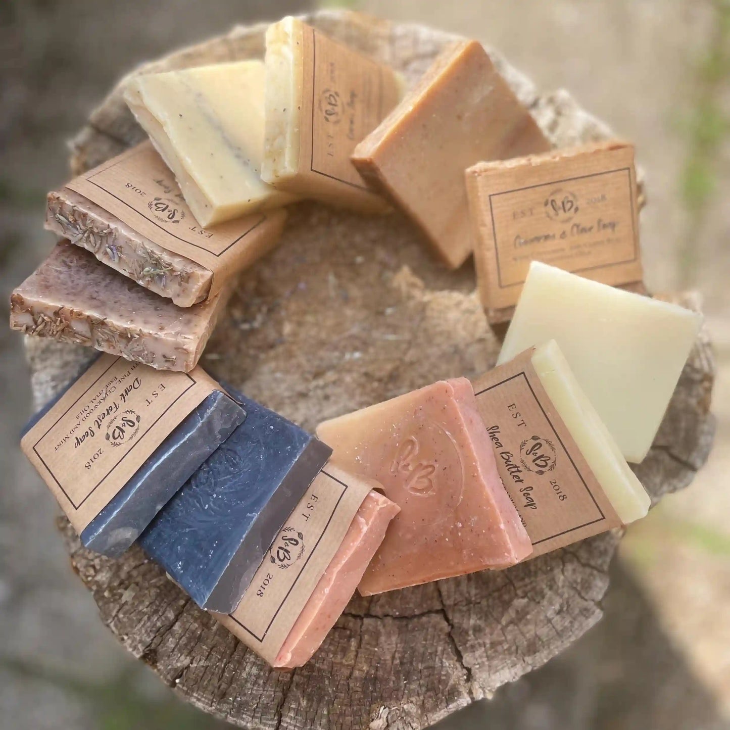 A collection of various handmade artisan soaps from Soapy & Bubbly UK, arranged in a circle on a rustic wood stump.