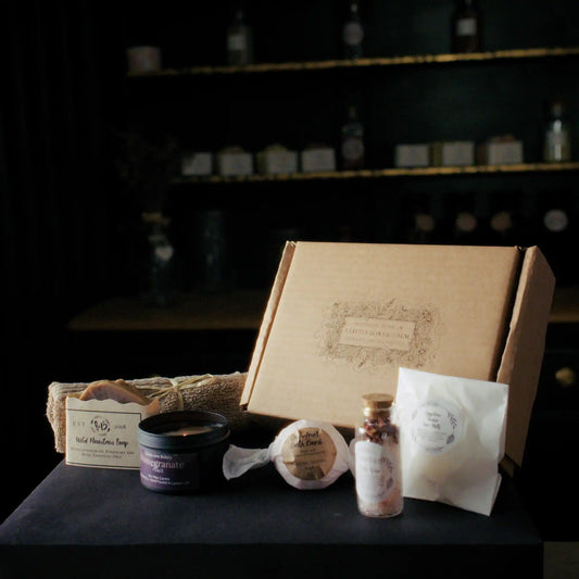 A_little_box_of_calm_02: A cardboard gift box with a floral label reading A Little Box of Calm displayed on a black surface alongside wellness items including a Wild Mountain Soap bar, a Pomegranate Noir candle, a bath bomb, bath salts, a wax melt package, and a rolled towel.