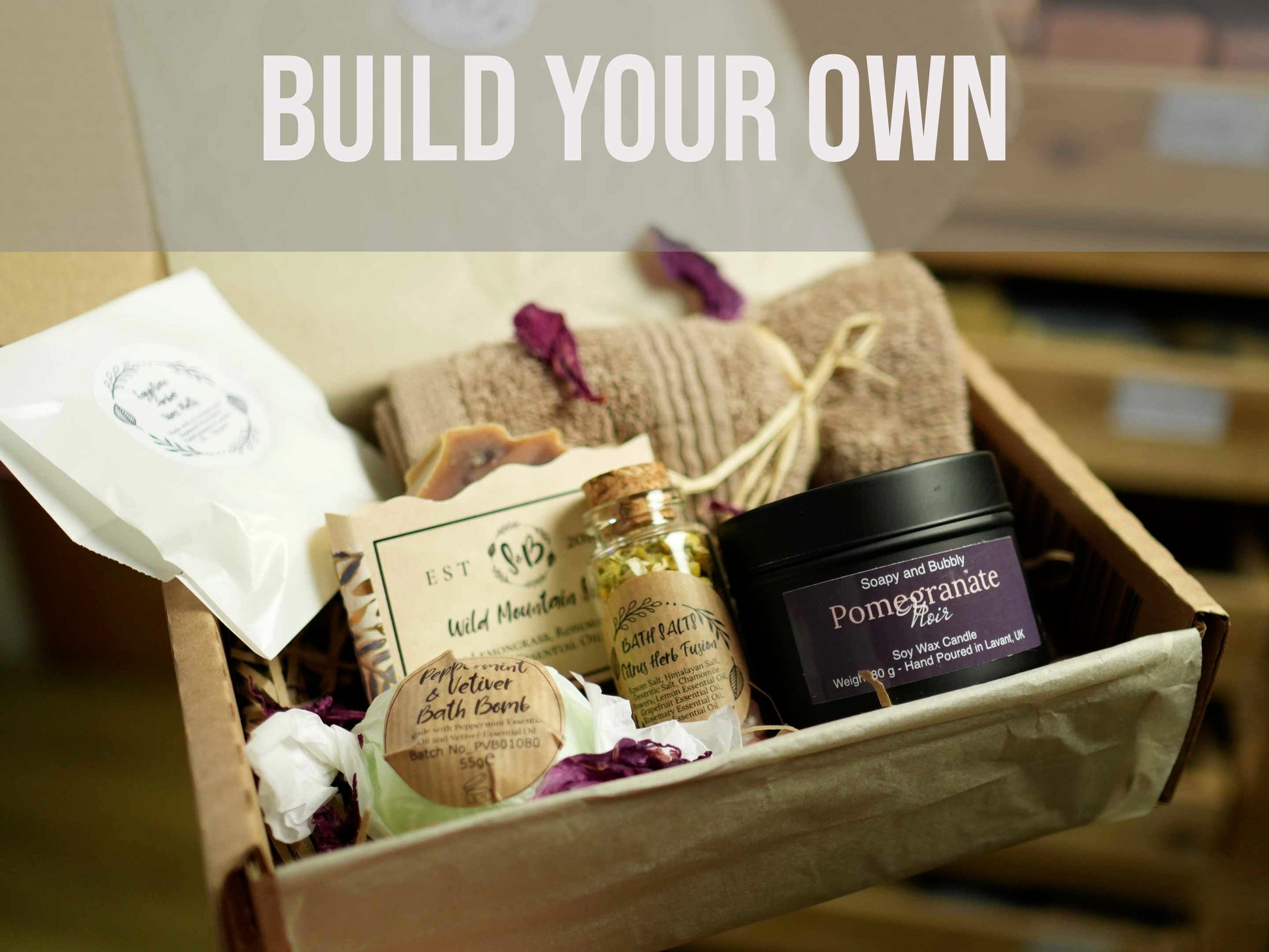 A Soapy and Bubbly open gift box containing a Pomegranate Noir soy wax candle, a Peppermint and Vetiver bath bomb, a glass jar of Citrus Herb Fusion bath salts, a Wild Mountain bar of soap, and a rolled brown towel.