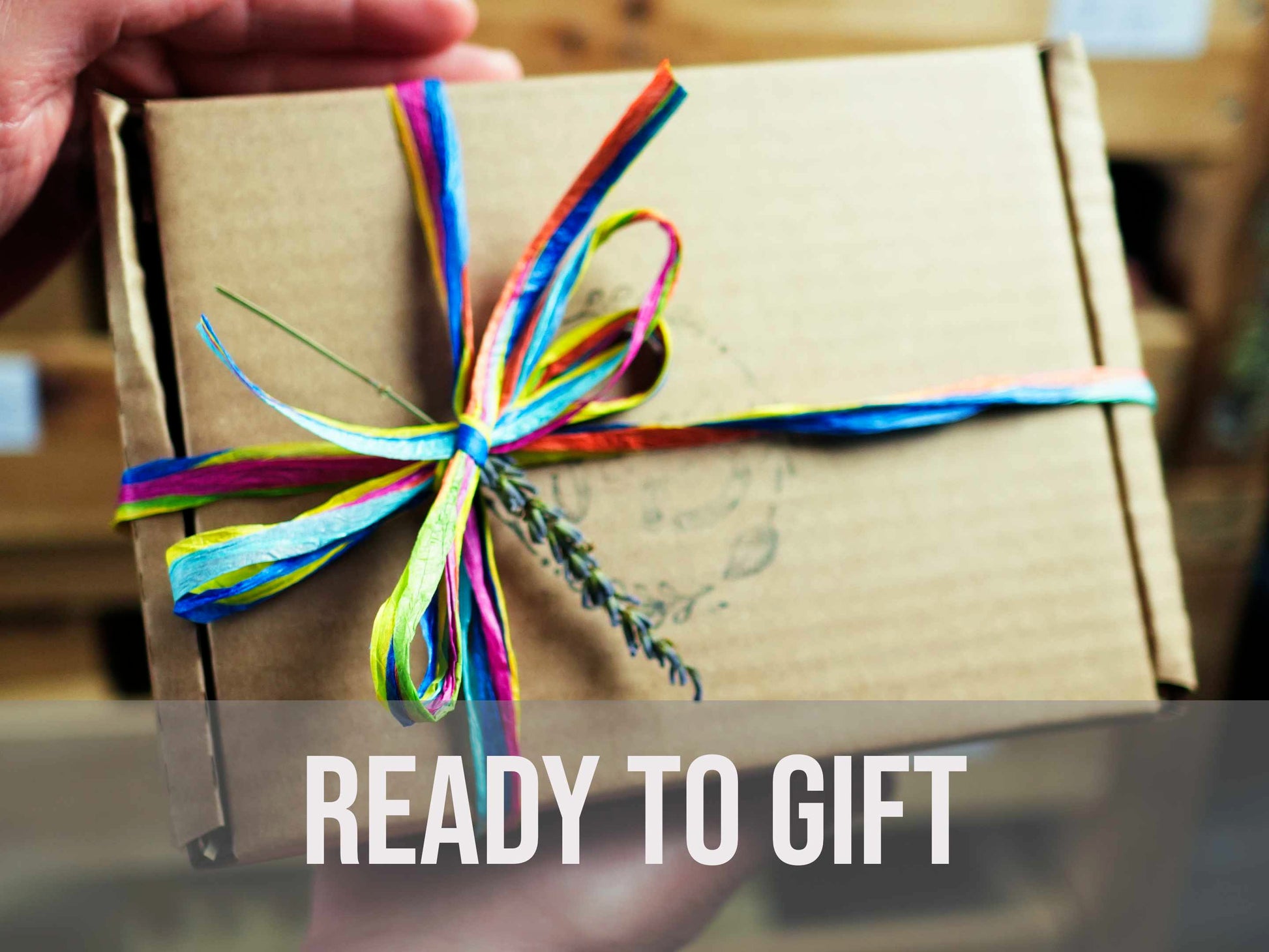 A person holding a closed brown cardboard gift box tied with a vibrant multi-colored ribbon and a sprig of dried lavender.