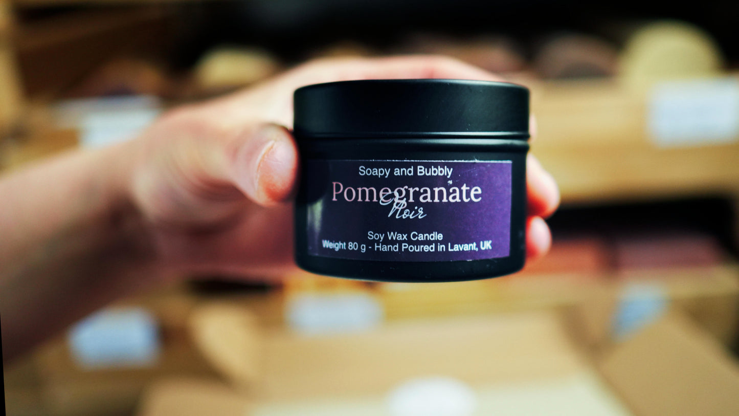 A person holding a black tin of Soapy and Bubbly Pomegranate Noir soy wax candle against a blurred background of wooden shelves.