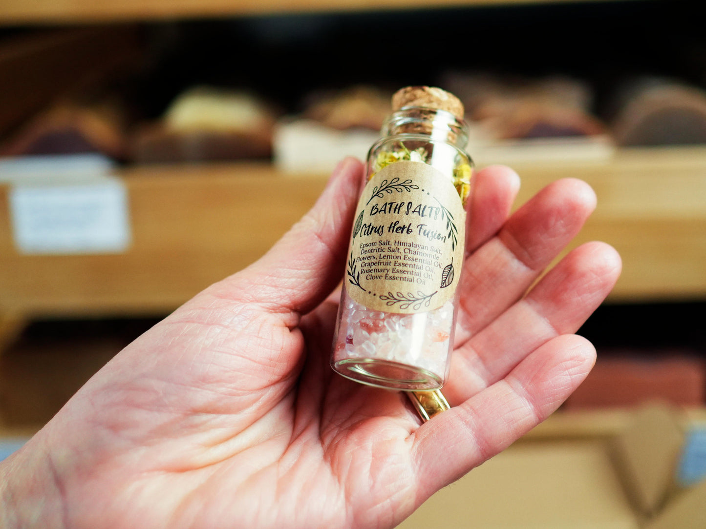 A hand holding a small glass bottle of Citrus Herb Fusion bath salts from Soapy and Bubbly with a cork stopper and a kraft paper label.
