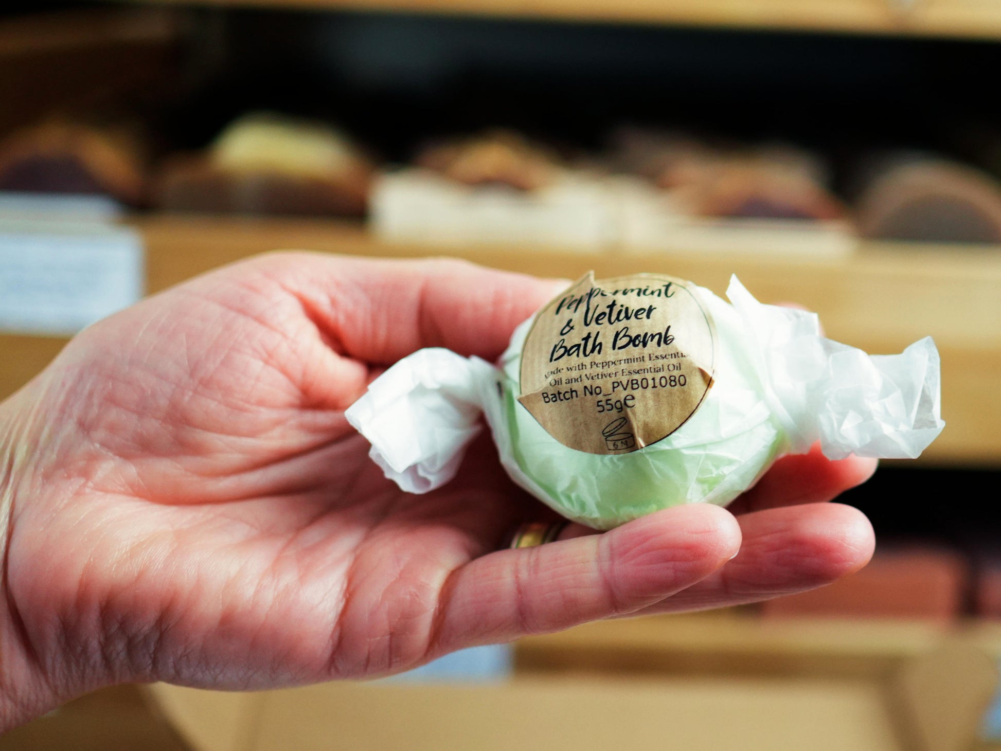 A hand holding a Soapy and Bubbly Peppermint and Vetiver Bath Bomb wrapped in white tissue paper with a brown circular label.