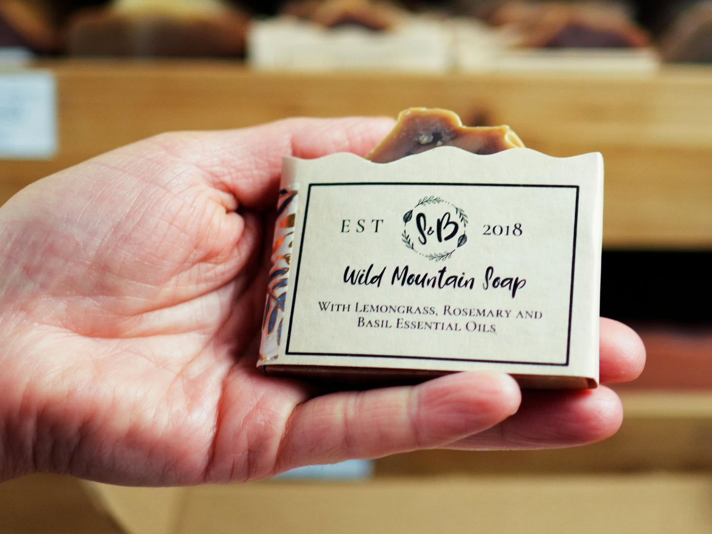A hand holding a bar of Wild Mountain Soap  from Soapy and Bubbly with Lemongrass, Rosemary and Basil Essential Oils in a cream paper wrapper.