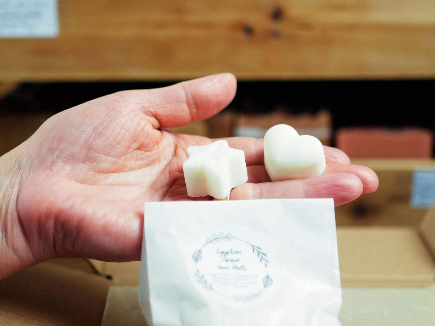 A hand holding a star-shaped and a heart-shaped white wax melt from Soapy and Bubbly above a small white paper bag labeled Egyptian Amber Wax Melts.
