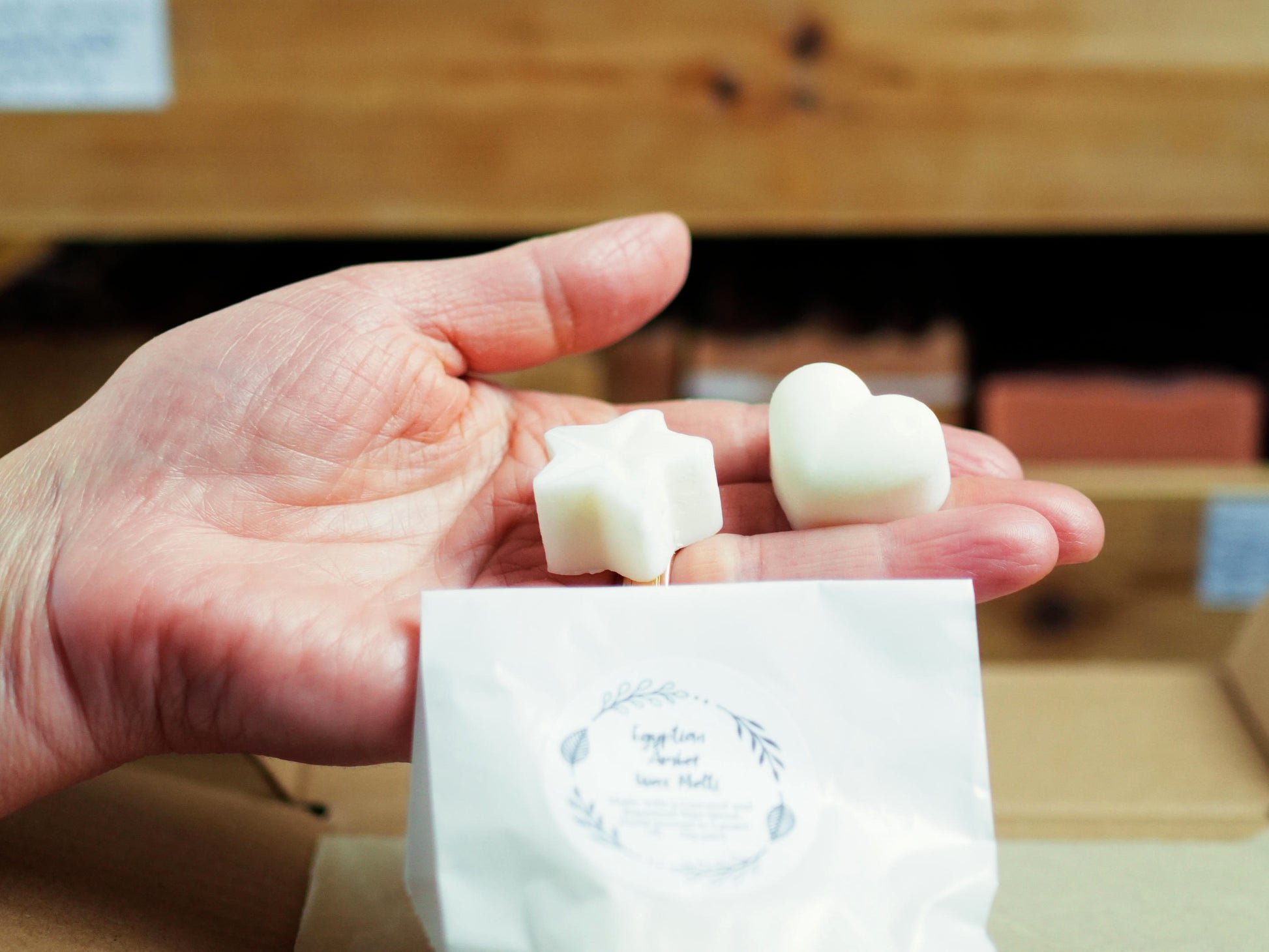 A hand holding a star-shaped and a heart-shaped white wax melt from Soapy and Bubbly above a small white paper bag labeled Egyptian Amber Wax Melts.