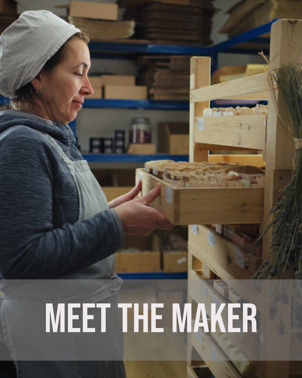 Mihaela, the Soapy and Bubbly owner wearing an apron and headcover sliding a wooden tray filled with handmade soaps into a shelving unit with a text overlay that reads MEET THE MAKER.