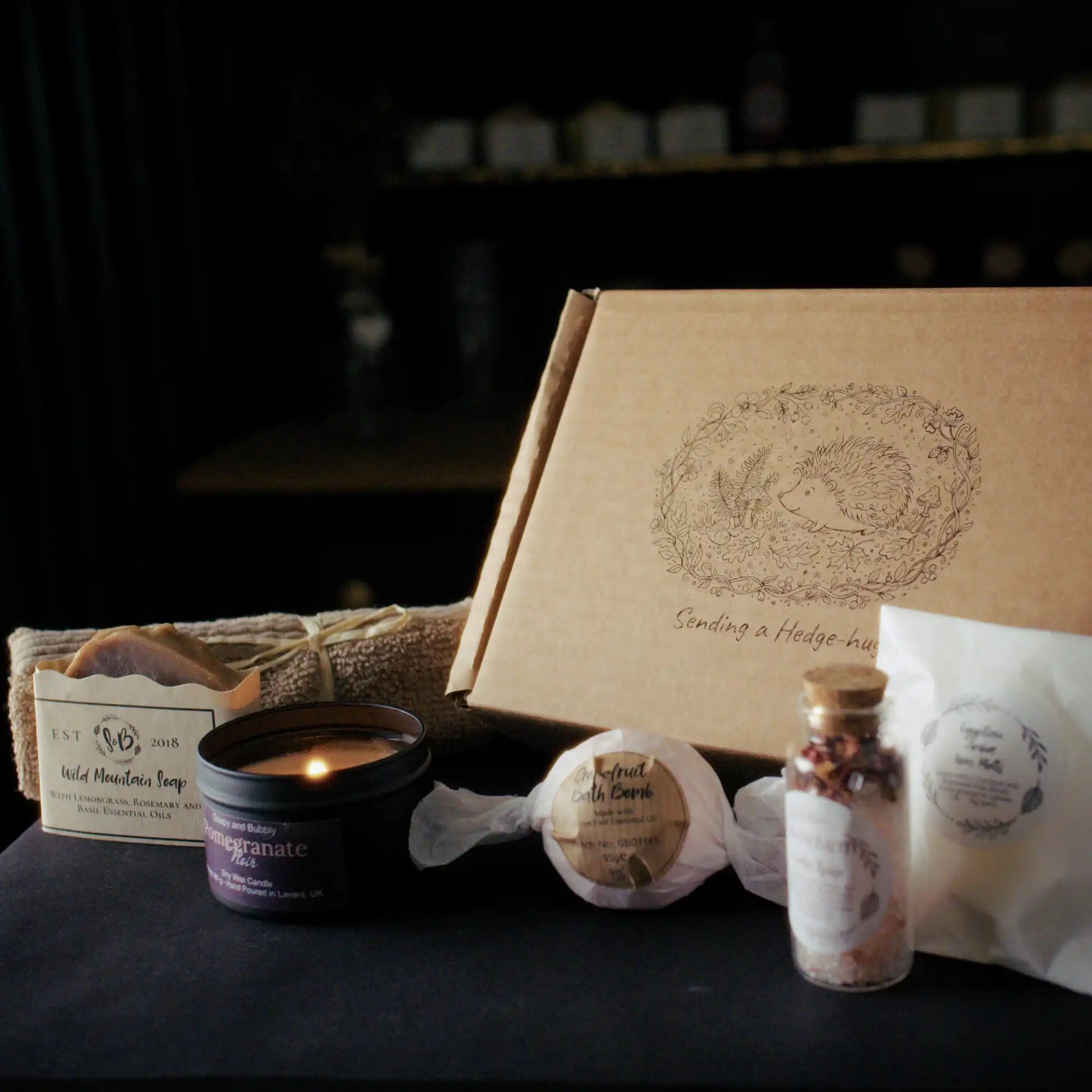 Sending_a_hedge_hug_01: A brown cardboard gift box with an illustrated hedgehog design and the text Sending a Hedge-hug, surrounded by self-care products including a lit candle, a Wild Mountain Soap bar, a bath bomb, a bottle of bath salts, and wax melts.